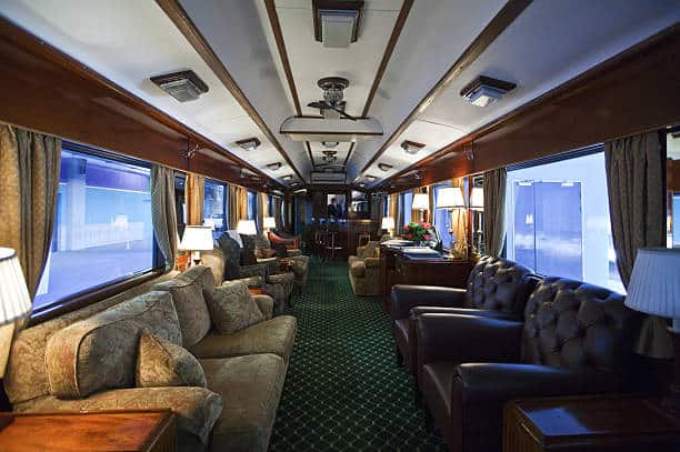 Luxurious train car interior with plush armchairs, sofas, and tables arranged in a cozy setting. The decor features wood paneling, large windows, and elegant lighting.