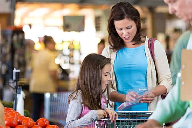 A woman and a girl stand near a grocery cart with vegetables. The woman holds a wallet while the girl looks at the cart.