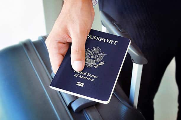 A person holds a United States passport next to a rolling suitcase handle.