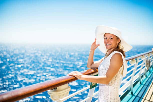 A woman in a white hat and dress stands by a cruise ship railing, smiling with the ocean in the background.