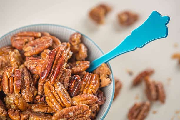 A bowl of roasted pecans with a blue spoon. Some pecans are scattered on the table.