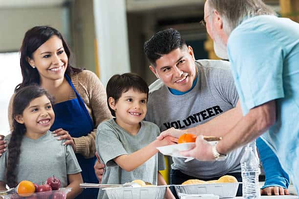 Volunteers serve food to a family at a community event. A young boy smiles while receiving a plate from an older man.