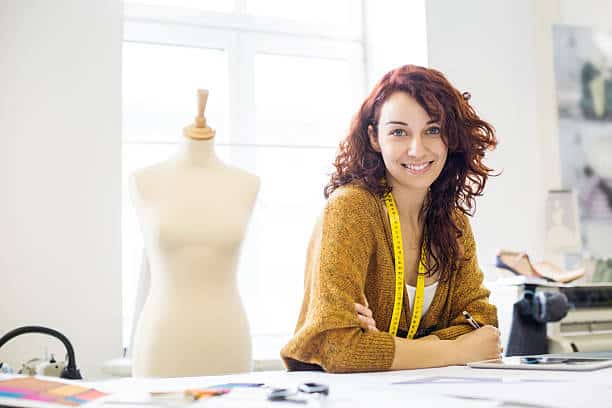 A woman with curly hair, wearing a yellow sweater and a measuring tape around her neck, sits at a desk in a design studio with a mannequin in the background.