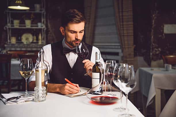 A man in formal attire sits at a restaurant table, tasting red wine and taking notes. Nearby, bottles of wine and glasses are arranged on the table.