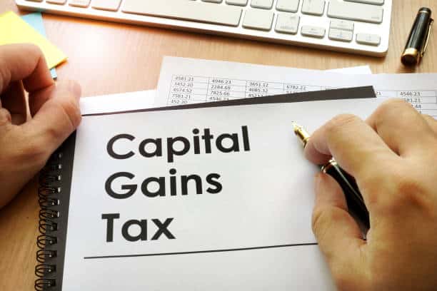 A person holds a document labeled "Capital Gains Tax" on a desk with financial papers, a pen, and a computer keyboard.