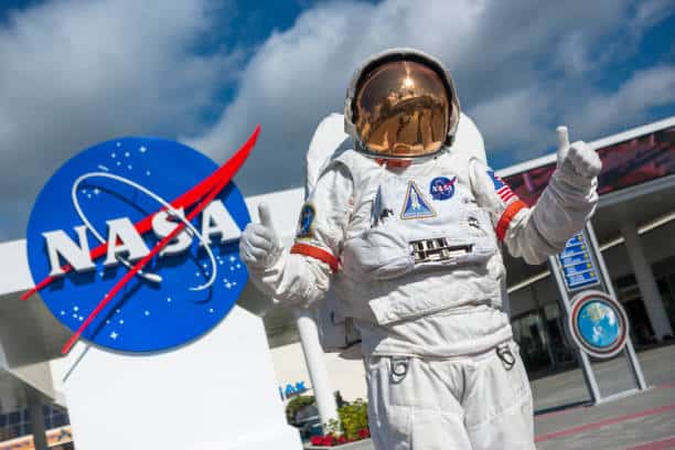 An astronaut in a spacesuit gives two thumbs up in front of a large NASA logo outside a building under a cloudy sky.