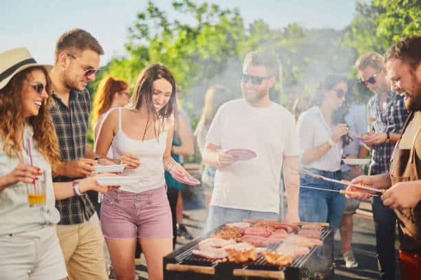 A group of people grilling and socializing outdoors on a sunny day. Some hold plates and drinks, while others chat around a barbecue with food cooking.