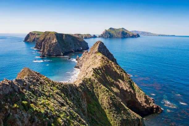 Aerial view of rugged cliffs extending into the ocean under a clear blue sky, with rocky coastlines and calm waters surrounding the land.