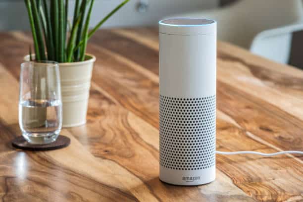A white smart speaker, resembling an Amazon Echo, on a wooden table next to a glass of water and a potted plant.