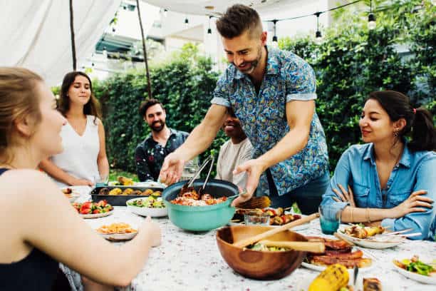 A group of people enjoying a meal outdoors around a table. One person is serving food from a pot, surrounded by various dishes. The setting is casual and relaxed.