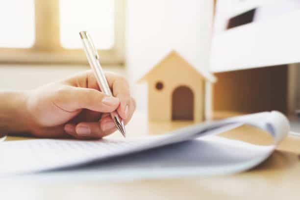 Hand holding a pen writing on paper, with a small wooden house model in the background.