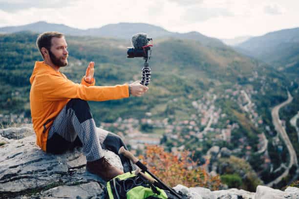 A man in an orange jacket sits on a rocky ledge, holding a camera on a flexible tripod. A scenic view of a valley and hills is in the background.