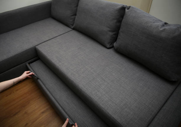 Person pulling out the bottom section of a gray sectional sofa on a wooden floor.