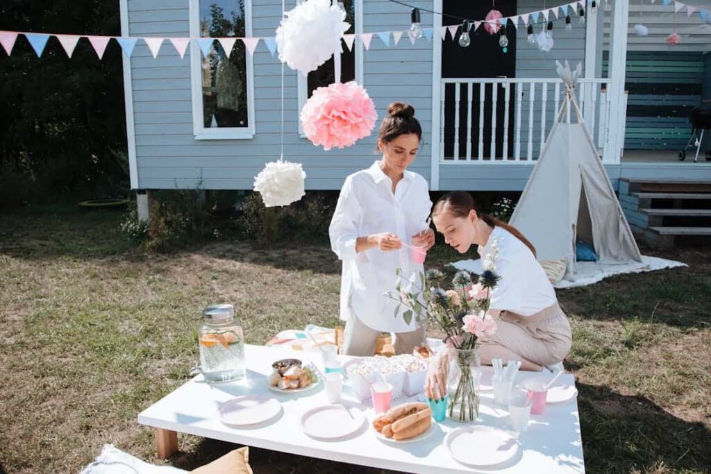 Two women preparing a table for an outdoor party with snacks, drinks, and flowers, in front of a house and teepee tent. Hanging decorations are visible above them.