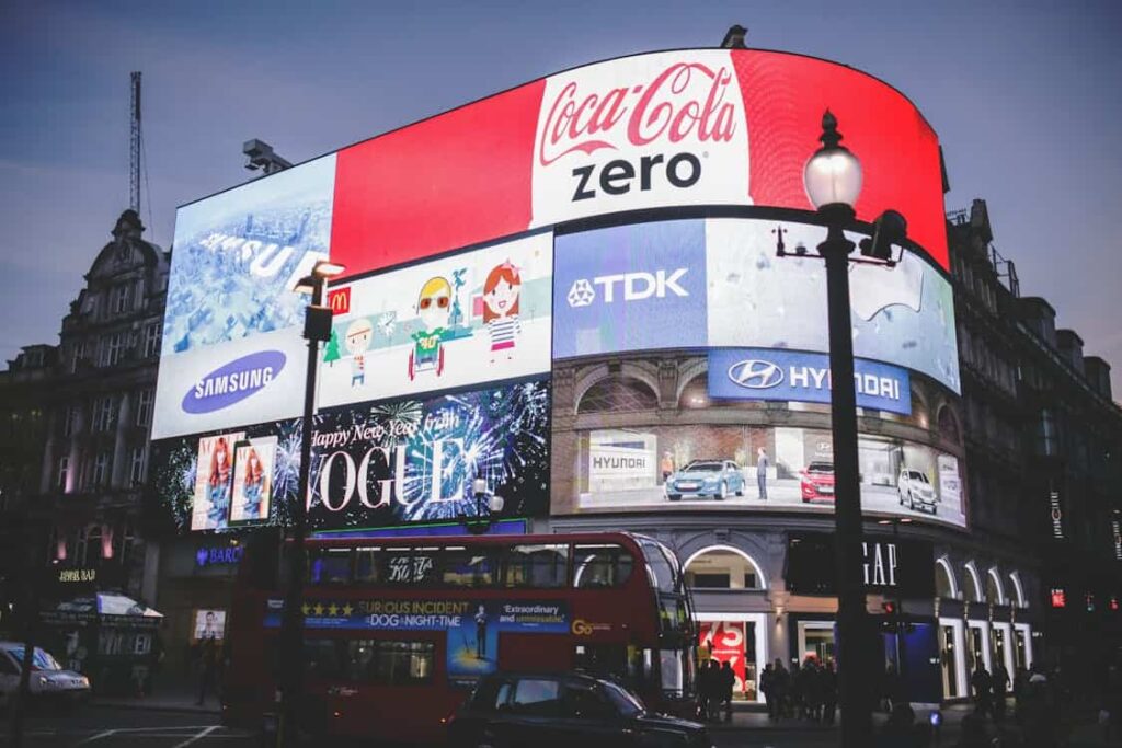 Bright digital billboards display various ads, including Coca-Cola Zero and Samsung, above a busy street featuring a red double-decker bus during dusk.