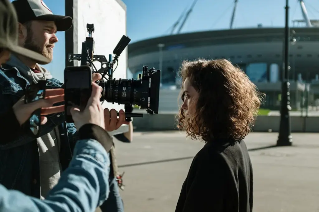A woman with curly hair is being filmed by a man with a camera rig. Another person holds a smartphone, capturing behind-the-scenes footage. A large stadium is visible in the background.
