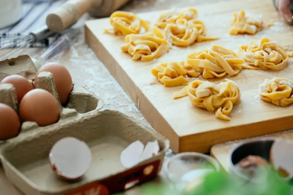 Fresh pasta nests on a floured wooden board beside brown eggs in a carton, with cracked shells nearby.