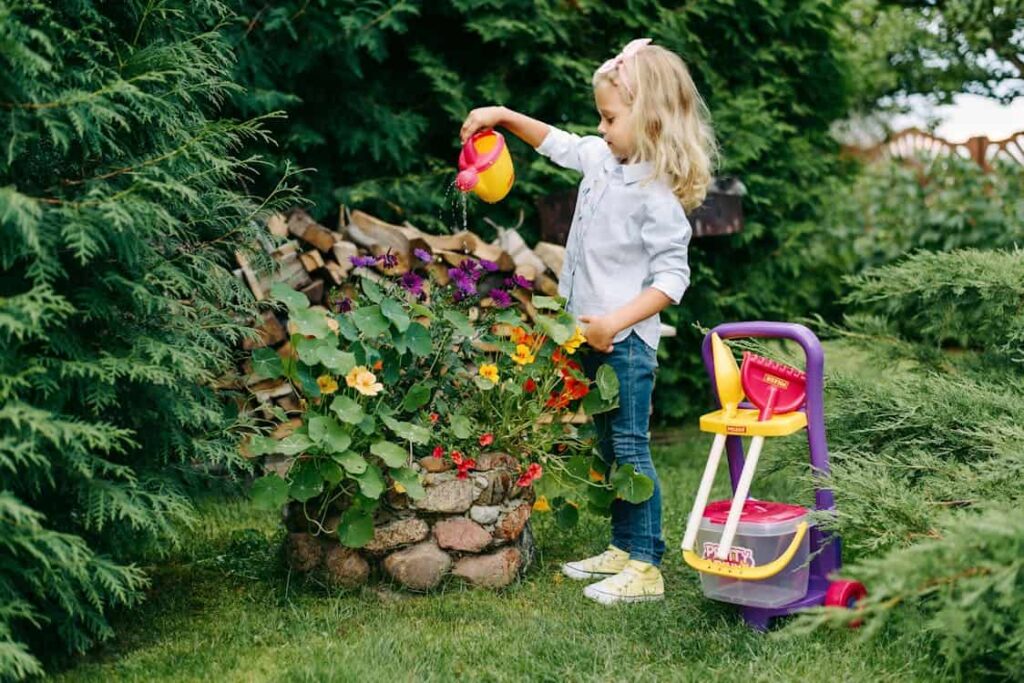 A young girl waters colorful flowers near a stone planter in a garden, with a toy vacuum beside her.