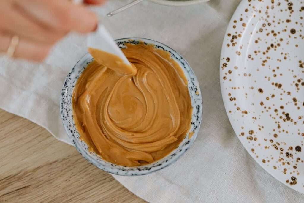 A bowl of smooth, creamy peanut butter with a person holding a white spatula. A partially visible plate with gold patterns is on the side.