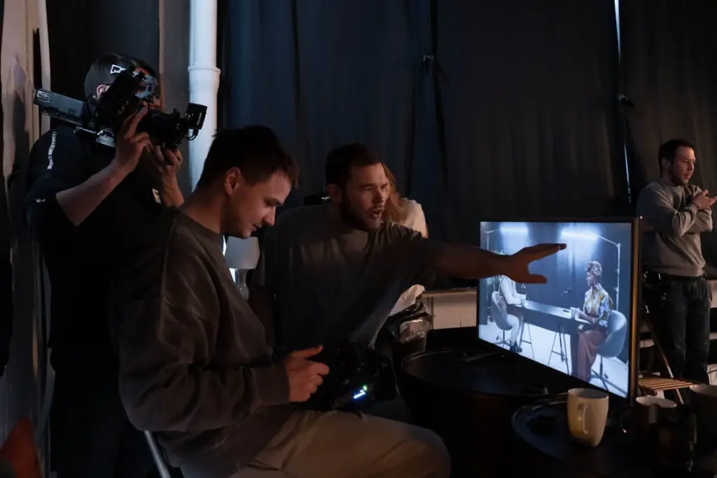 People in a dimly lit room working with camera equipment and a monitor displaying a scene. One person gestures towards the screen.