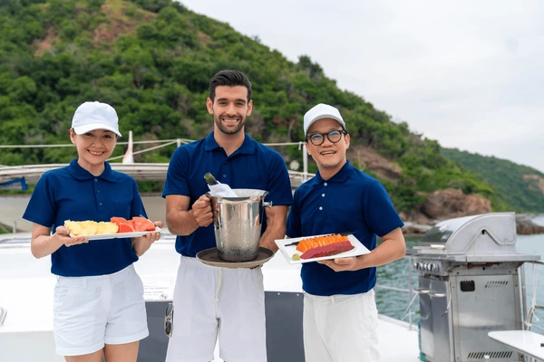 Three people in blue polo shirts and white shorts stand on a yacht, holding plates of food and a champagne bottle in an ice bucket, with a forested hill and sea in the background.