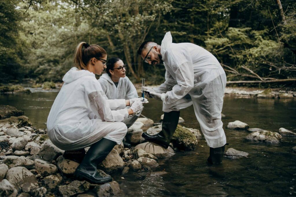 Three people in protective suits and boots collect water samples in a forested stream area.