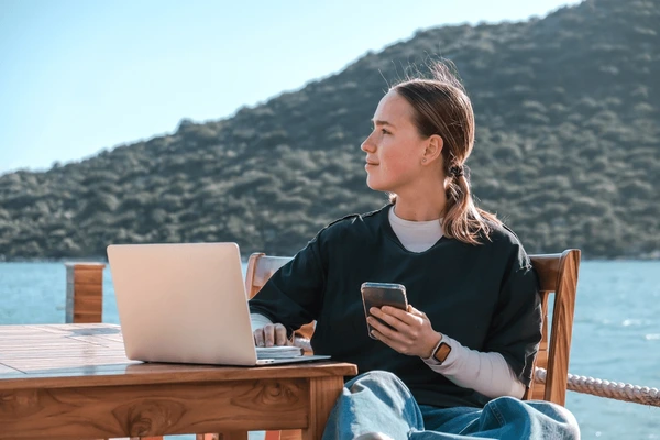 A person sitting at a table outdoors with a laptop and smartphone, looking towards the water and hills in the background.