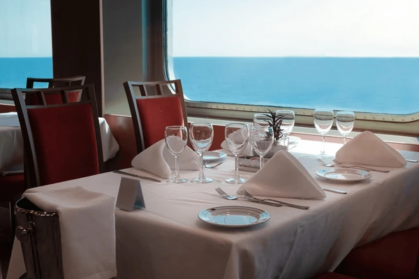 Dining table set for four with white tablecloth, napkins, glasses, and cutlery, next to a window revealing an ocean view.