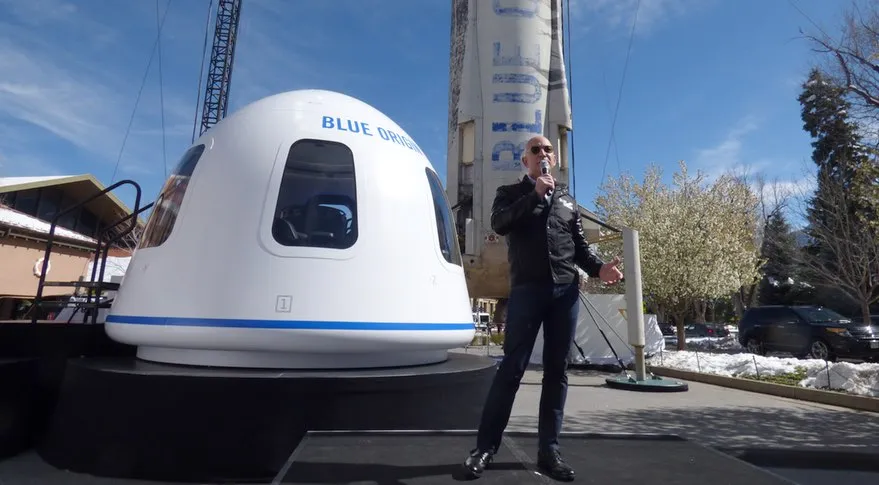 A person speaks into a microphone near a Blue Origin space module on display with a rocket in the background, under a clear sky.