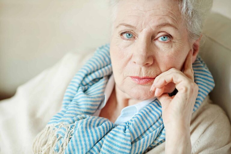Elderly person with short gray hair wearing a blue-striped scarf, gazes intently while resting their head on their hand.