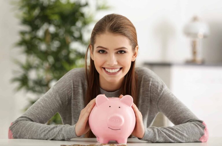 Woman smiling, leaning on a pink piggy bank at a table, with a plant and lamp in the background.