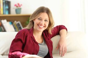 Woman with long hair wearing a red shirt and gray top, sitting on a white sofa, smiling. Bookshelf and potted plant visible in the background.