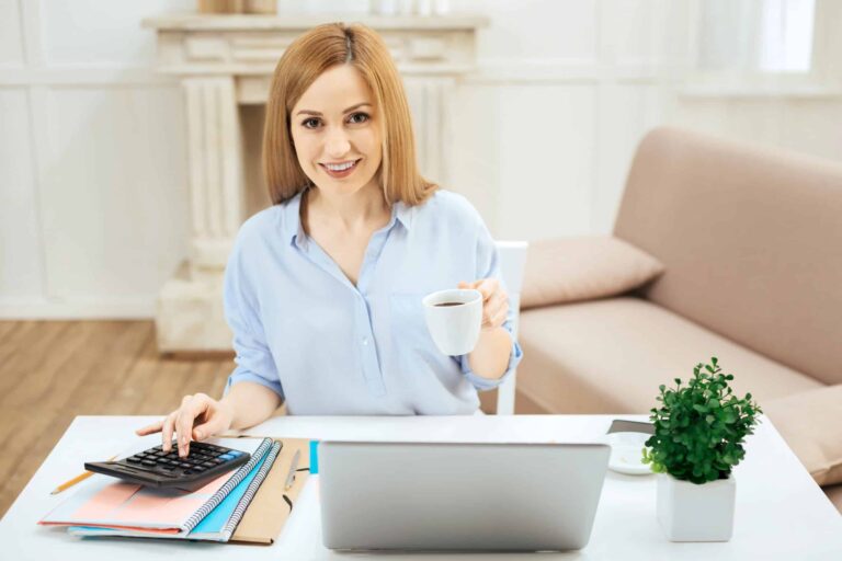 Woman sitting at a desk with a laptop, holding a cup of coffee and using a calculator, with notebooks and a small plant nearby.
