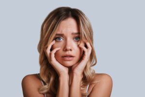 A concerned woman with blonde hair holds her face with both hands, looking towards the camera against a light gray background.