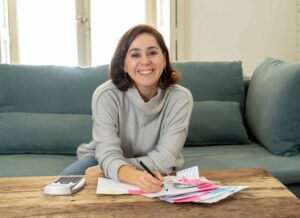 Person smiling, seated on a sofa, writing on papers with a pen. A calculator is nearby on a wooden table. Bright room with natural light from windows in the background.