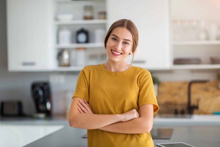 Person in a yellow shirt standing in a kitchen with arms crossed, smiling. White cabinets and kitchen items are in the background.
