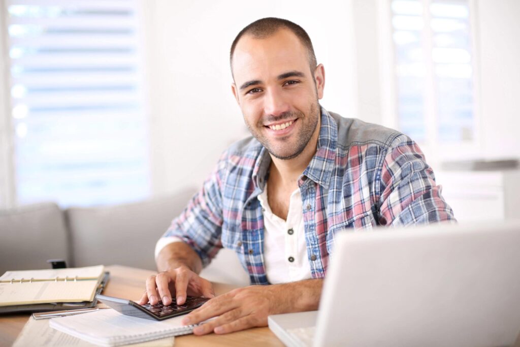 A person in a plaid shirt sits at a desk with a laptop, smiling at the camera.