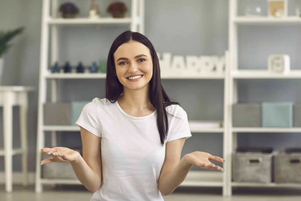 Woman with dark hair in a white t-shirt sits indoors, smiling and shrugging. Shelves with decorative items are in the background.