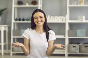 Woman with dark hair in a white t-shirt sits indoors, smiling and shrugging. Shelves with decorative items are in the background.