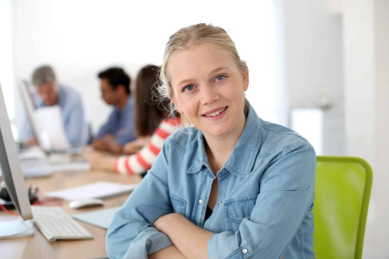 Person in a denim shirt sitting at a desk, smiling at the camera. Others work in the background.