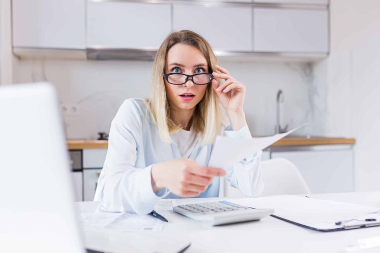 A surprised woman holding a document, adjusting her glasses, sits at a table with a calculator and papers in a bright room.