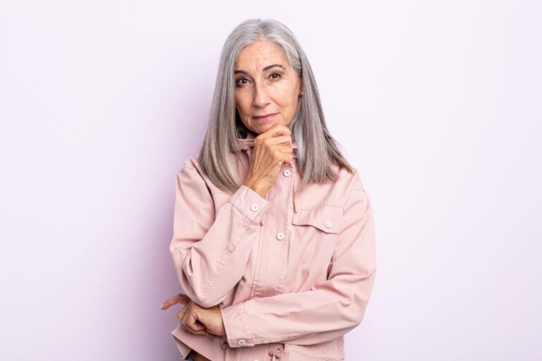 An older woman with long gray hair stands against a light-colored background, wearing a pink jacket and resting her chin on her hand.