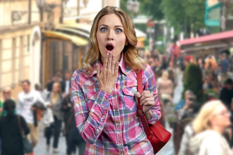 A woman in a plaid shirt stands in a busy street with a surprised expression, her hand covering her mouth. People walk in the blurred background.
