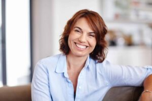 A woman with short brown hair smiles while seated on a sofa, wearing a light blue shirt. The background is softly blurred, suggesting an indoor setting.