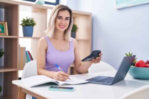 A woman sits at a table with a laptop, notebook, and smartphone, smiling while holding a calculator.