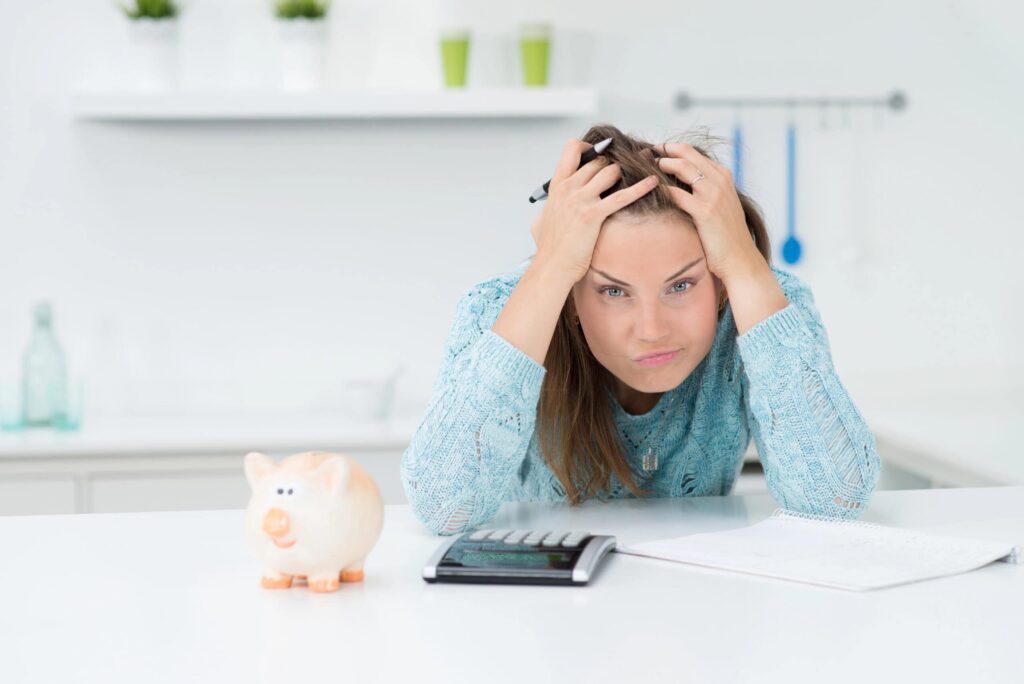 A frustrated person holds their head while sitting at a table with a piggy bank, calculator, and notepad in a bright room.