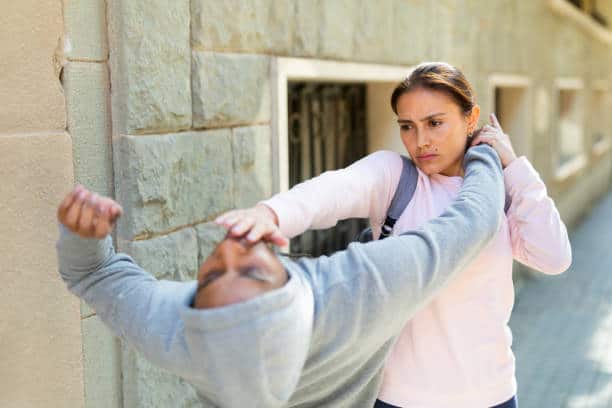 A woman in a pink hoodie defends herself by pushing away a man in a gray hoodie on a sidewalk with a brick wall.