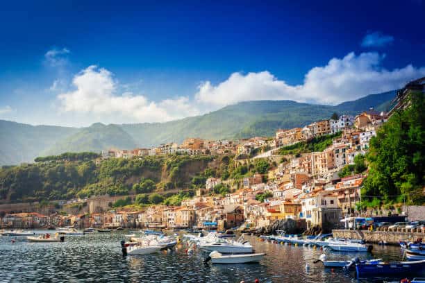 Coastal village with colorful buildings on a hillside, boats docked in the foreground, and greenery-covered mountains in the background under a blue sky with clouds.