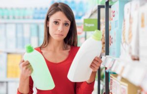 A woman in a red shirt stands in a store aisle holding and comparing two detergent bottles, one green and one white, with a thoughtful expression.