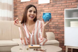 A woman sits on a couch, smiling, holding a blue piggy bank and a coin. Coins and a notepad with a pen are on the table in front of her. Brick wall in the background.